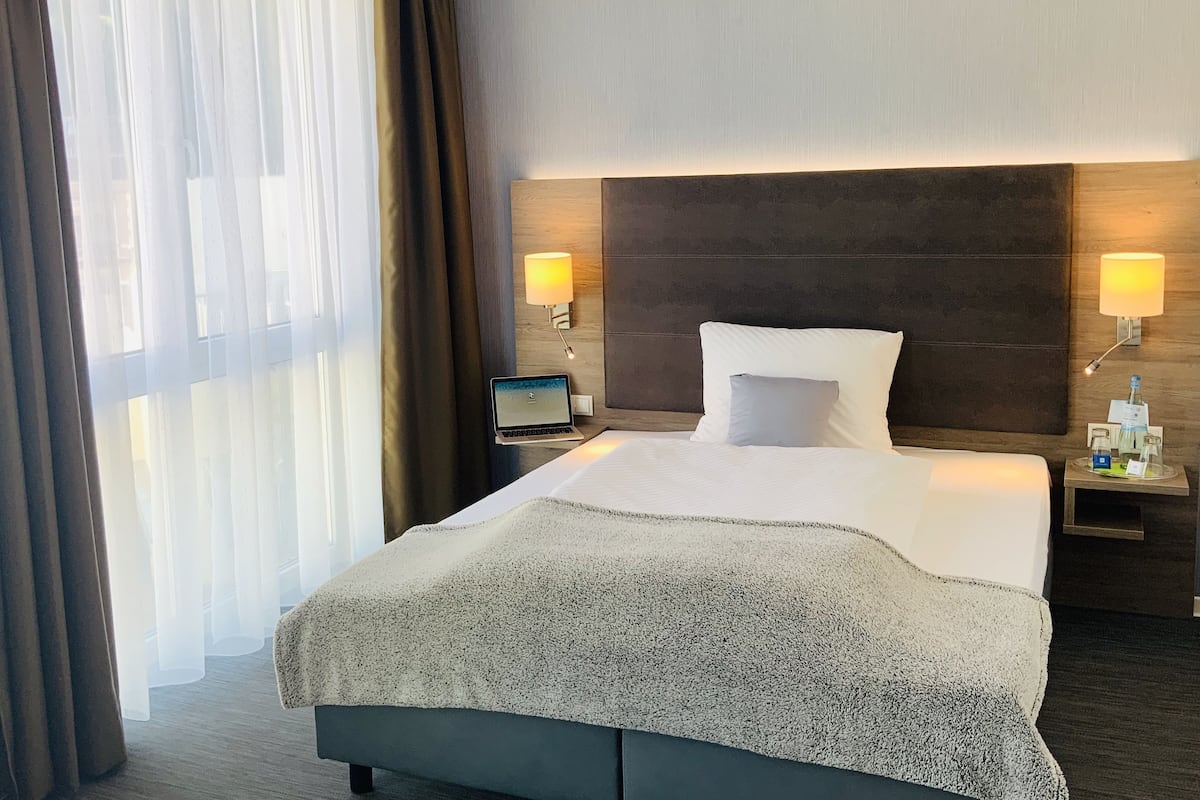 A hotel room with a bed, two bedside lamps, a laptop on the nightstand, and a window with curtains.