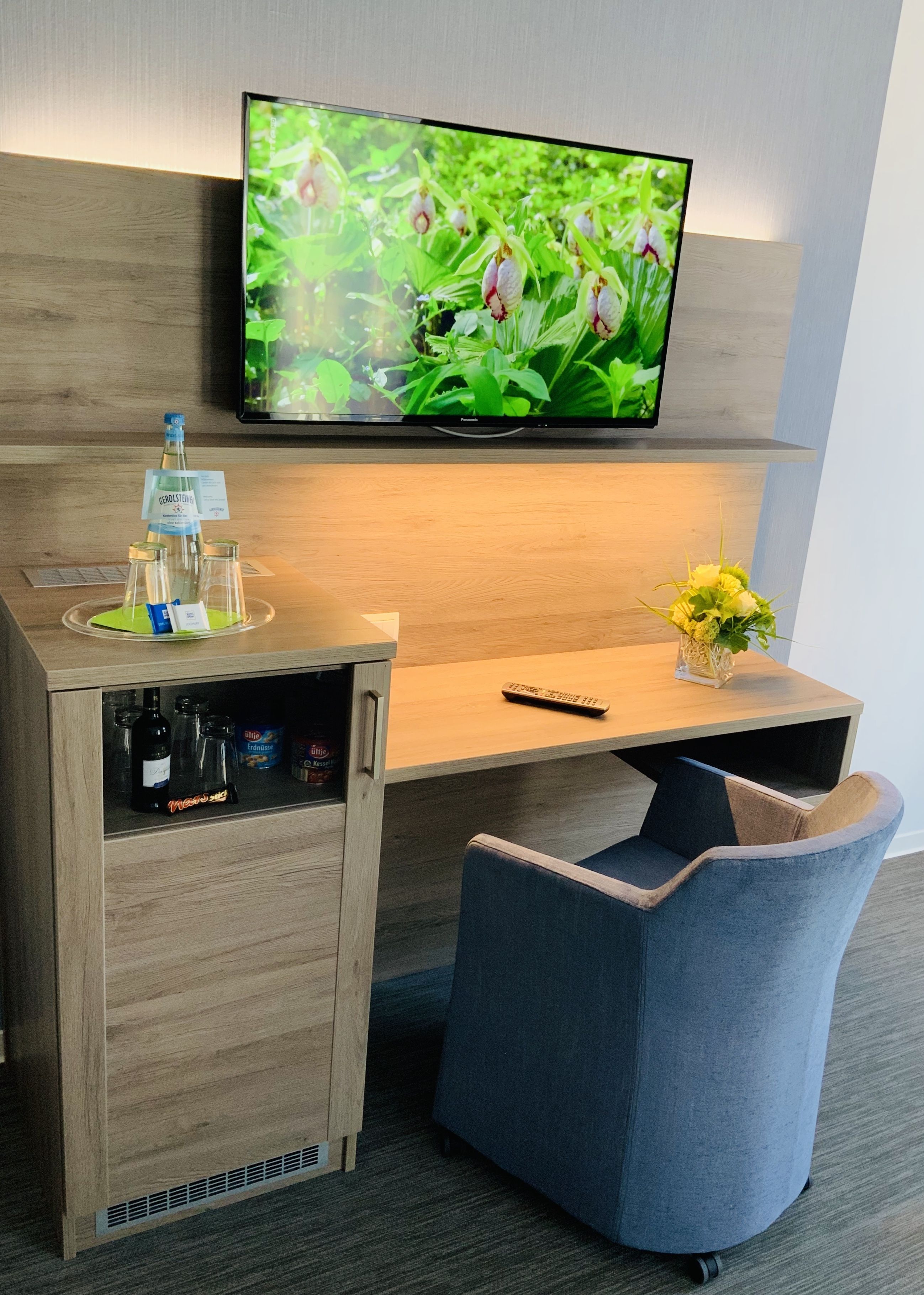 Ein Hotelzimmer mit einem Holztisch, einem an der Wand befestigten Fernseher, einer Fernbedienung, einer Vase mit Blumen und einem blauen Sessel.