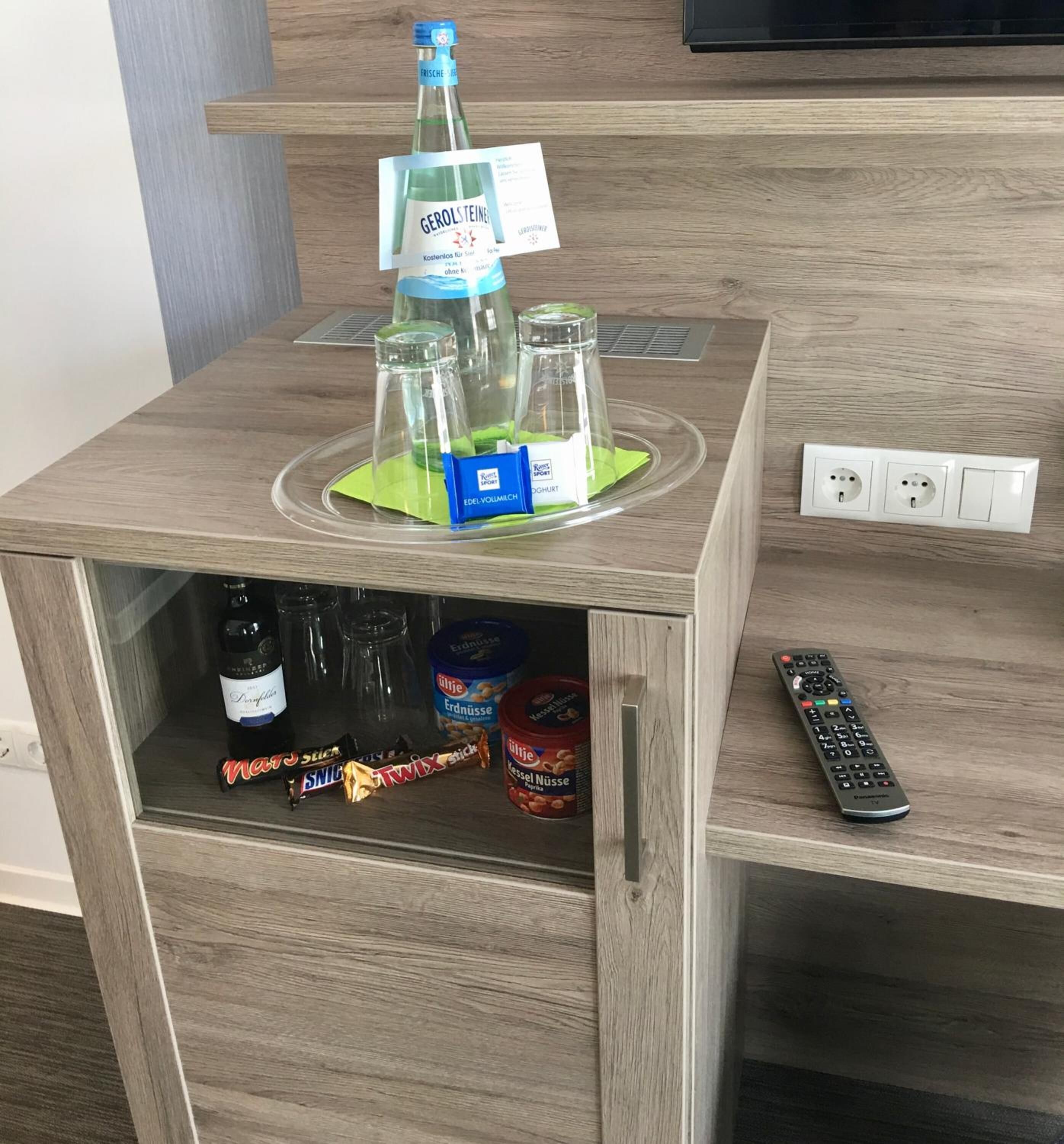 Eine Holzminibar mit einer Gerolsteiner-Flasche, zwei Gläsern, einem grünen Servietttuch und einer Fernbedienung.