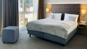 Quarto de hotel com uma cama grande, duas luminárias de cabeceira, uma mesinha de cabeceira pequena, um banquinho cinza e vista para o exterior por uma janela ampla.
