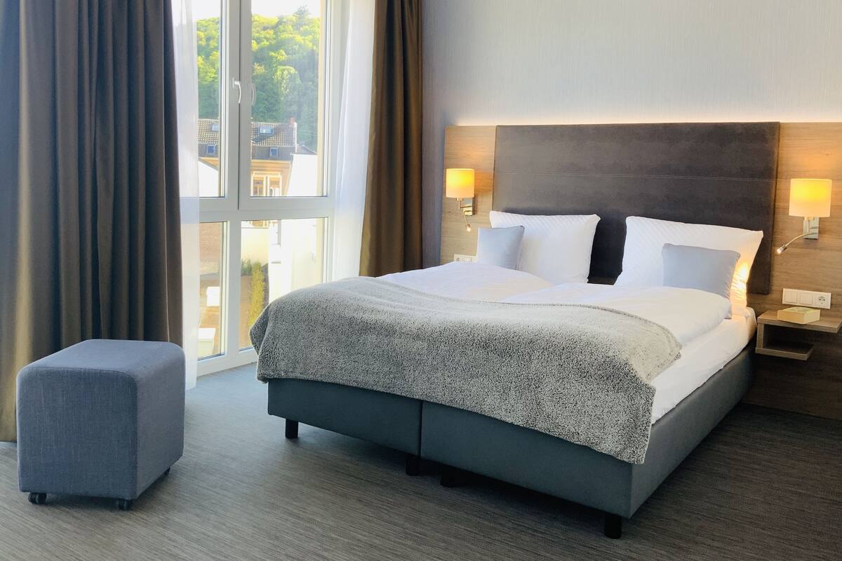 A hotel room with a large bed, a bedside table with a lamp, a small ottoman, and a view of the outside through a large window.