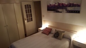 Une chambre à coucher comprenant un lit, deux tables de chevet, une commode, une porte avec une fenêtre et un tableau accroché au mur.