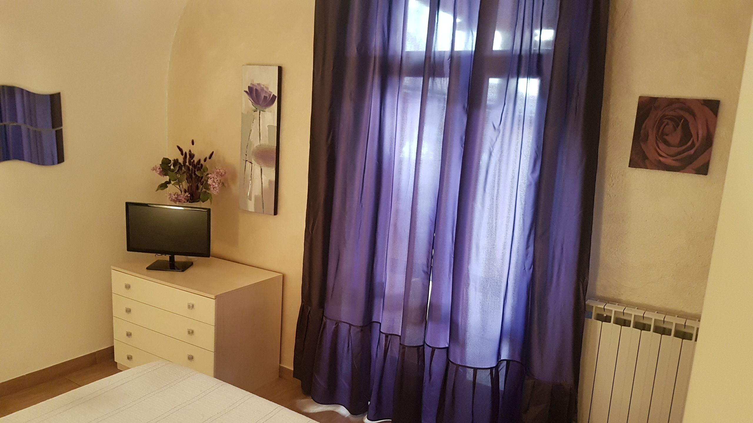 A bedroom with a bed, a TV on a stand, a dresser, a window with purple curtains, and a radiator.