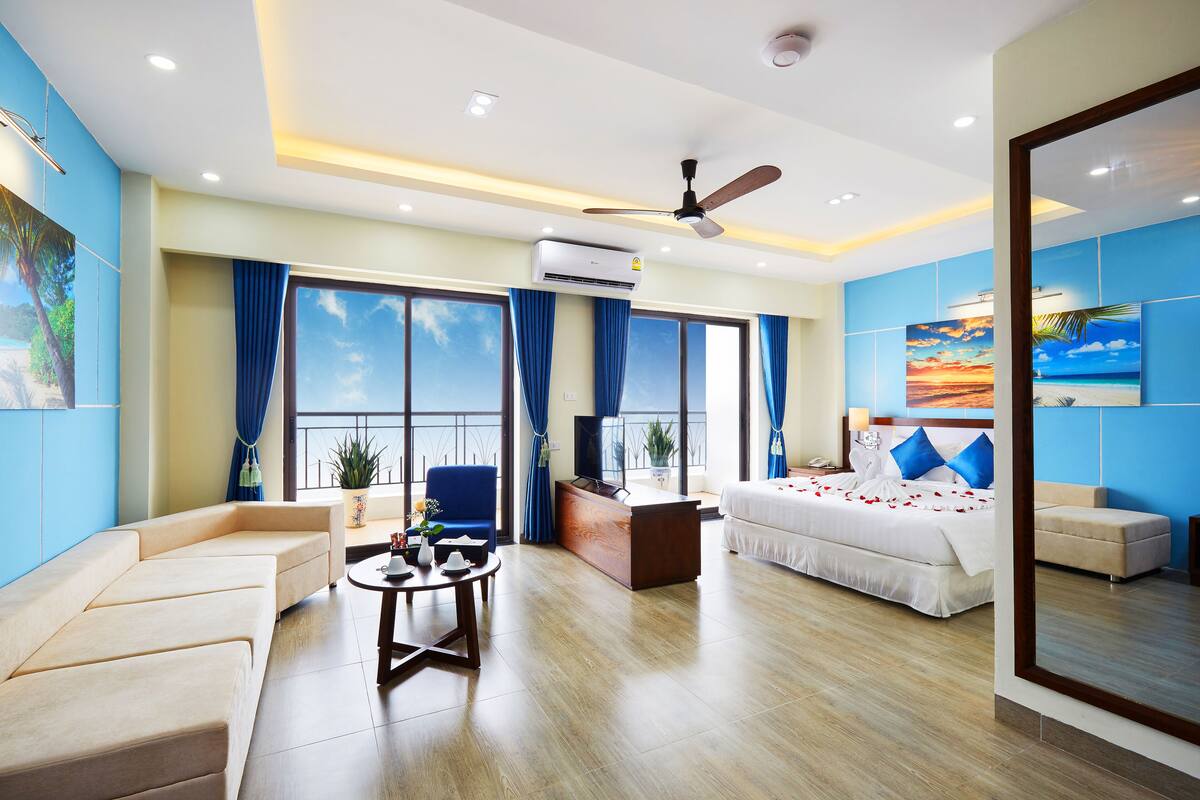 A modern hotel room with a large bed, a sofa, a desk, and a balcony with a view of the sky.