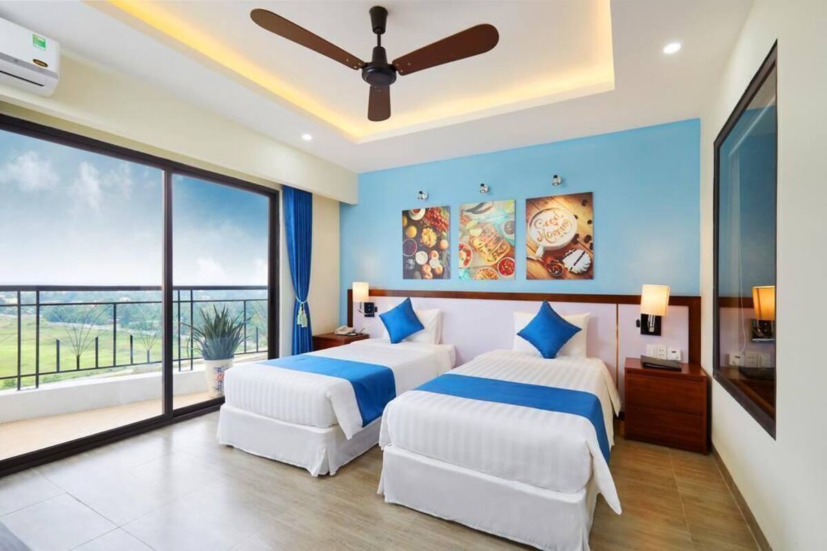 A hotel room with two beds, a ceiling fan, a large window with a view, and artwork on the wall.