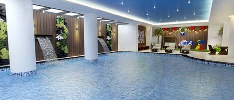 Amplia zona de piscina interior con suelo de baldosas azules, múltiples columnas y un mostrador de recepción con plantas decorativas y un mural colorido.