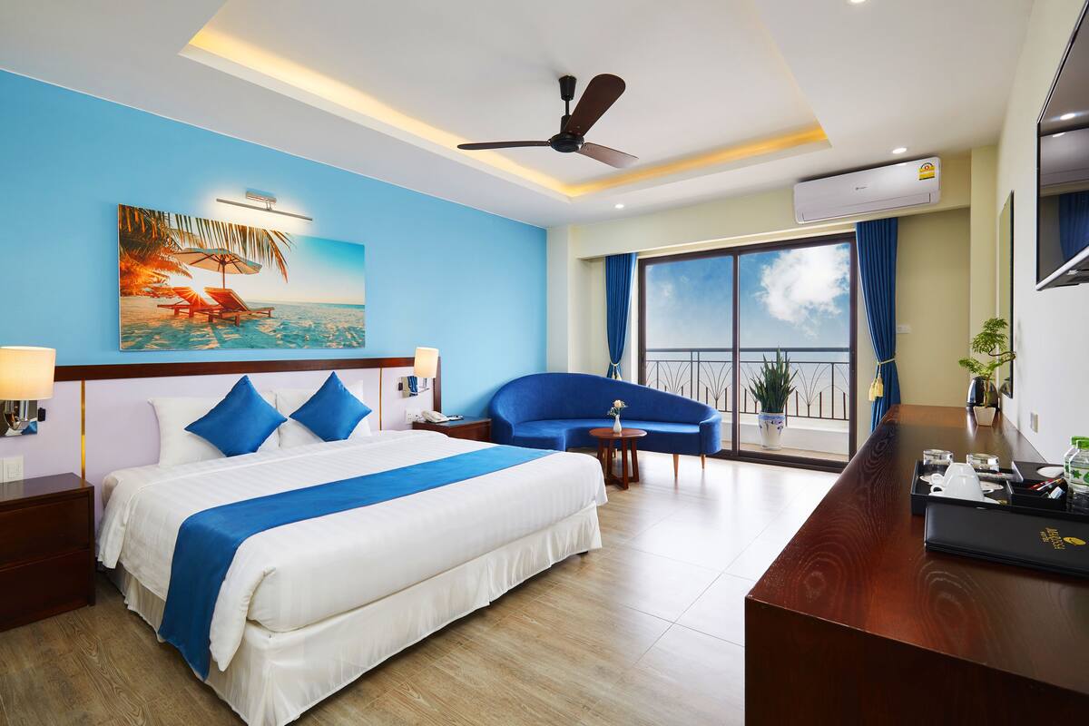 A hotel room with a large bed, a desk, a blue armchair, a balcony with a view, and an air conditioning unit.