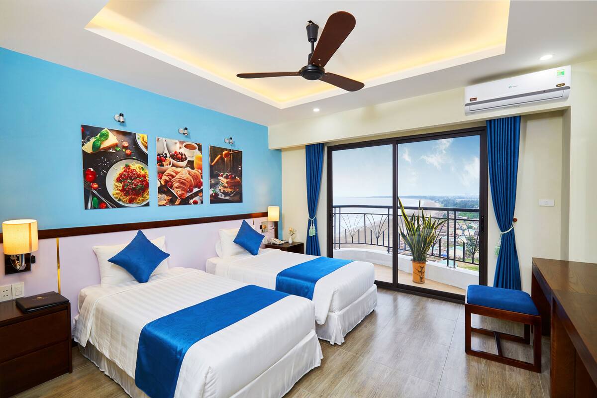A hotel room with two beds, a ceiling fan, a large window with a view, and a dining area with a table and chairs.