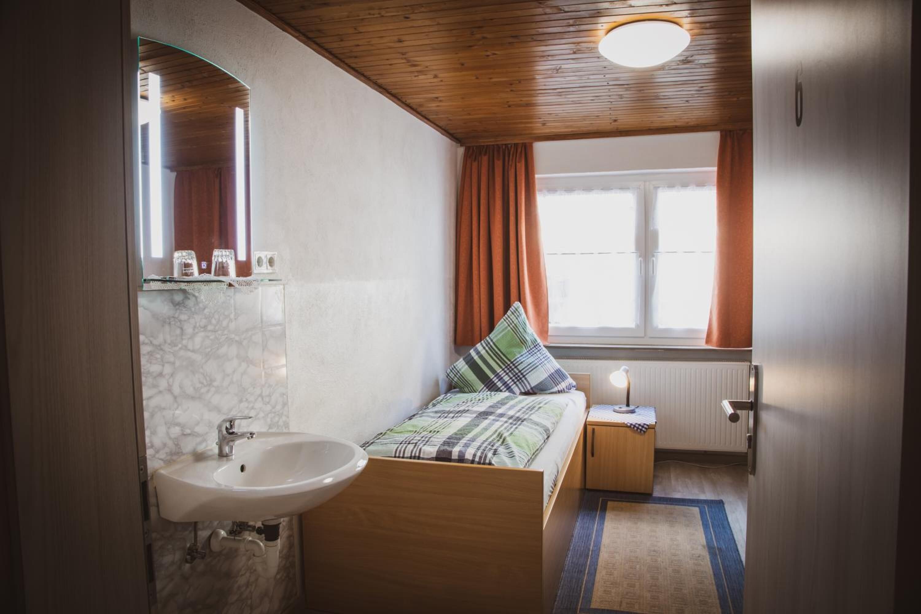 A compact bedroom with a single bed, a bedside table, a nightstand, a mirror, and a window with curtains.