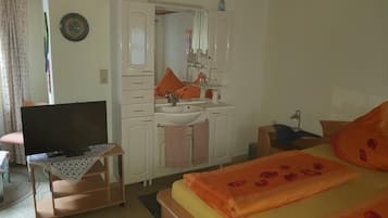Quarto com cama, televisão sobre suporte, cômoda com espelho e pia.