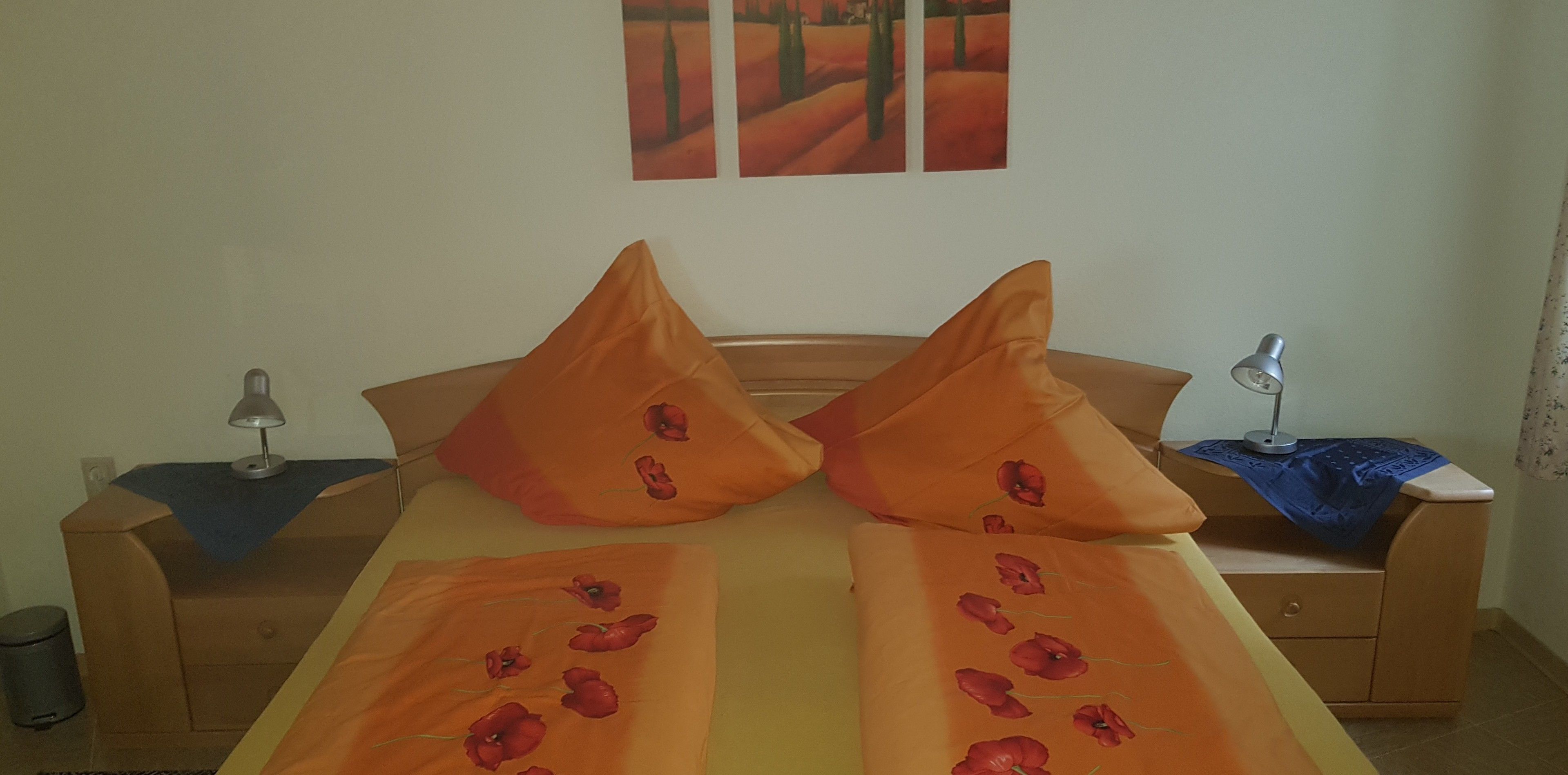 Un letto con due cuscini arancioni, un comodino con una lampada e un tavolino con lampada sul lato opposto.