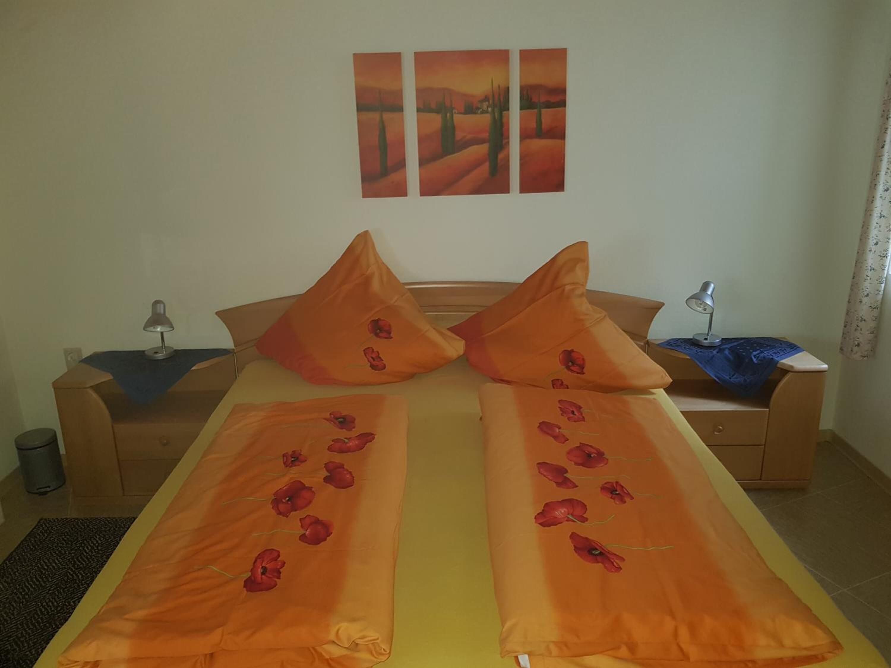 Una cama doble con ropa de cama naranja y diseños de amapolas, una mesita de noche con una lámpara y una pared decorada con tres cuadros.