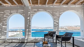 Terraza soleada con tres ventanales arqueados que dan a una vista costera, con una mesa, sillas y un vaso de agua.