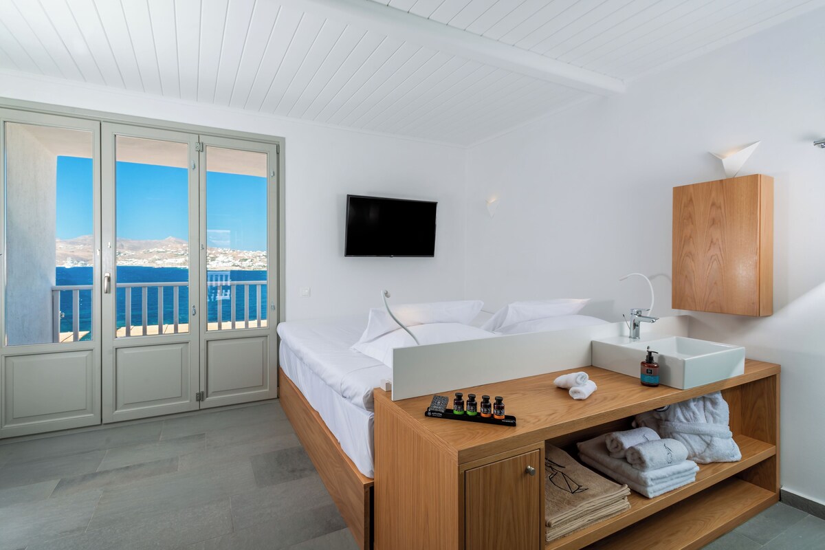 A hotel room with a bed, a TV, a wooden bedside table with toiletries, a sink, and a view of the sea.