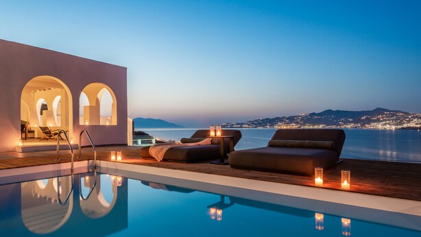 Zona junto a la piscina con sillones de descanso y velas, con vistas a una masa de agua y luces de la ciudad a lo lejos.