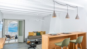 Un espacio habitable moderno con una zona de comedor de madera, un sofá con cojines, una mesita redonda con una taza y vistas al mar a través de una puerta abierta.