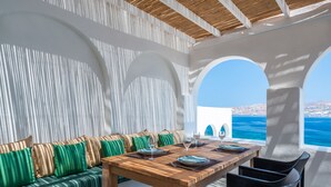 Un espacio de comedor al aire libre con luz solar, mesa de madera dispuesta para cuatro personas, sofá con cojines verdes y vista al mar.
