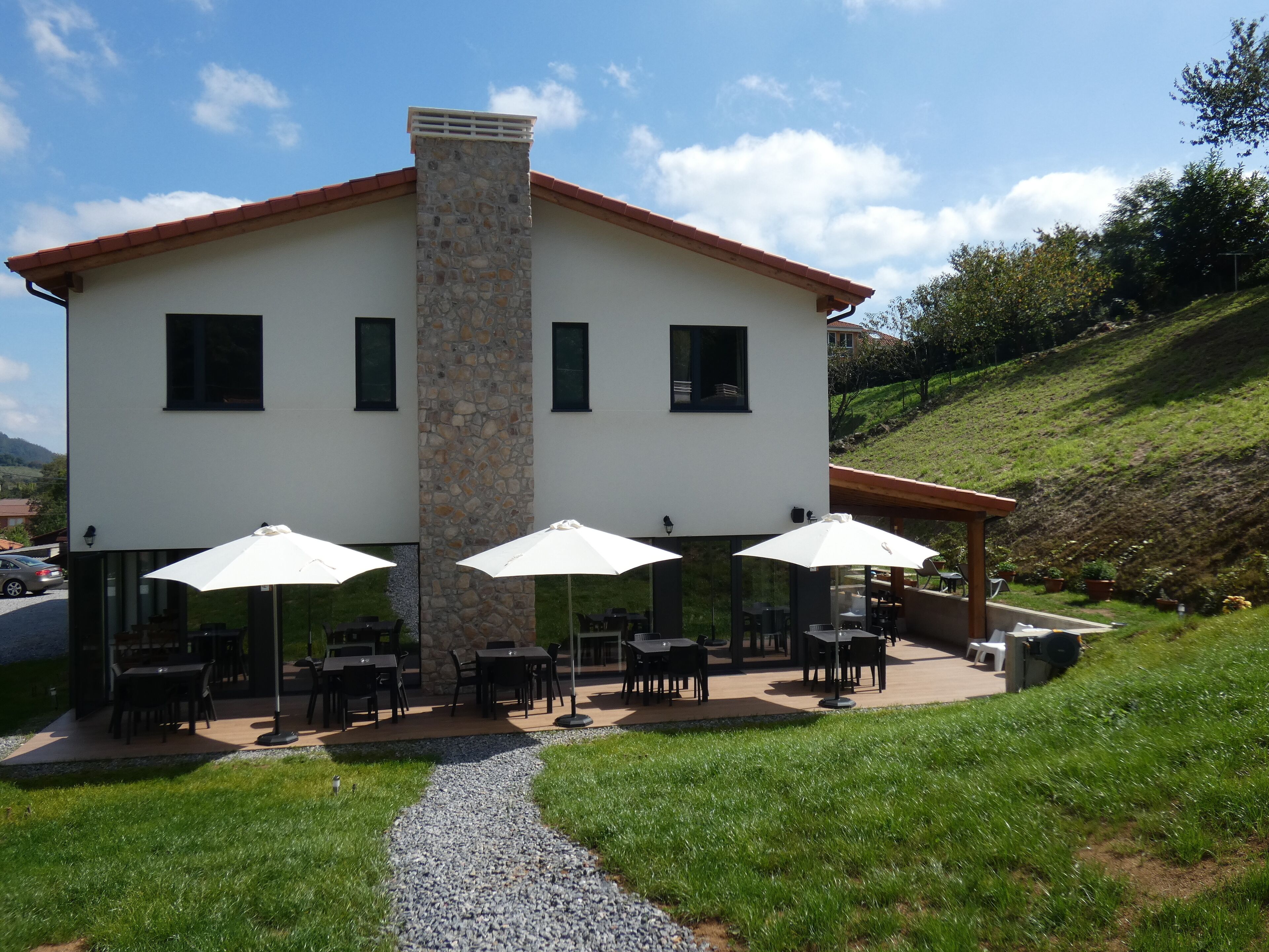 A two-story building with a stone chimney, outdoor seating with umbrellas, and a gravel path leading to the entrance.