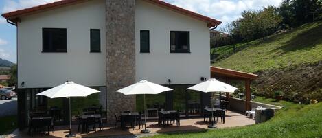 A two-story building with a stone chimney, outdoor seating with umbrellas, and a gravel path leading to the entrance.