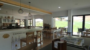 A modern dining area with wooden chairs and tables, a kitchen in the background, and a view of a grassy outdoor area through large windows. - Casa Sueño (Salas)