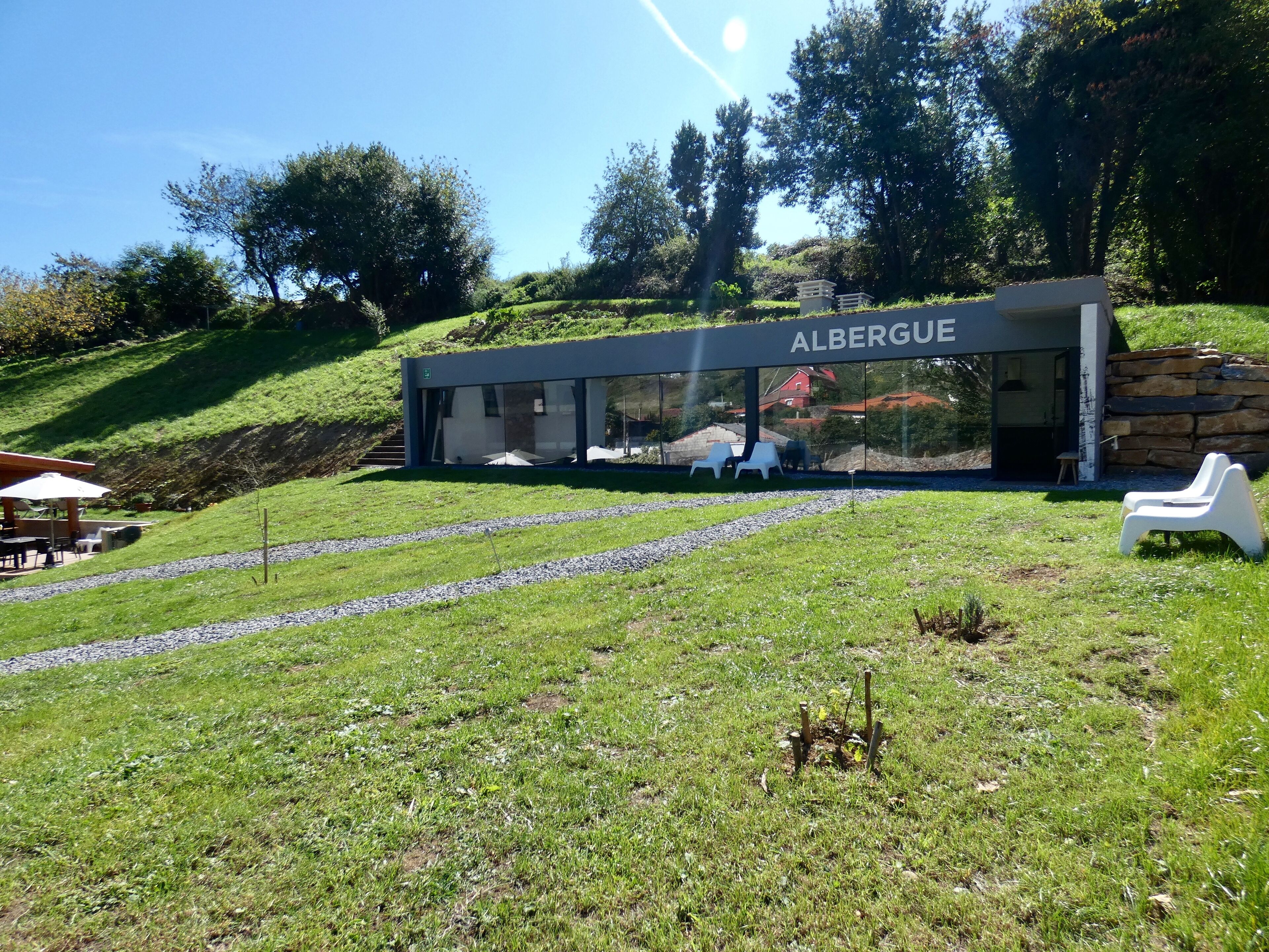 Un edificio moderno con el nombre 'ALBERGUE' en la fachada, rodeado de un área verde con algunas sillas de plástico blancas y un muro de piedra.