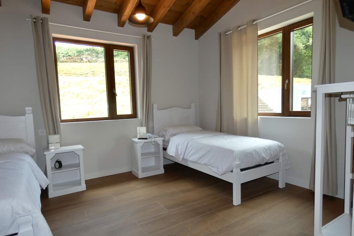 A bedroom with a wooden ceiling, two beds, a bedside table with a lamp, and a window with curtains.