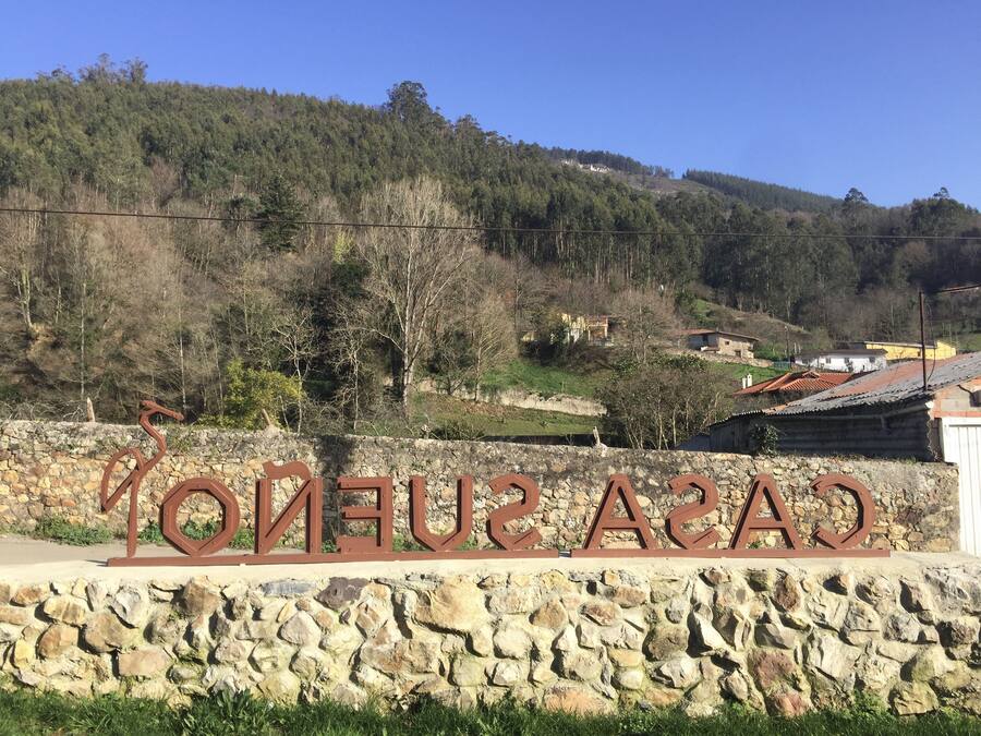 Un muro de piedra con el letrero "CASA SENERO" y una vista de un pueblo con casas y árboles al fondo.