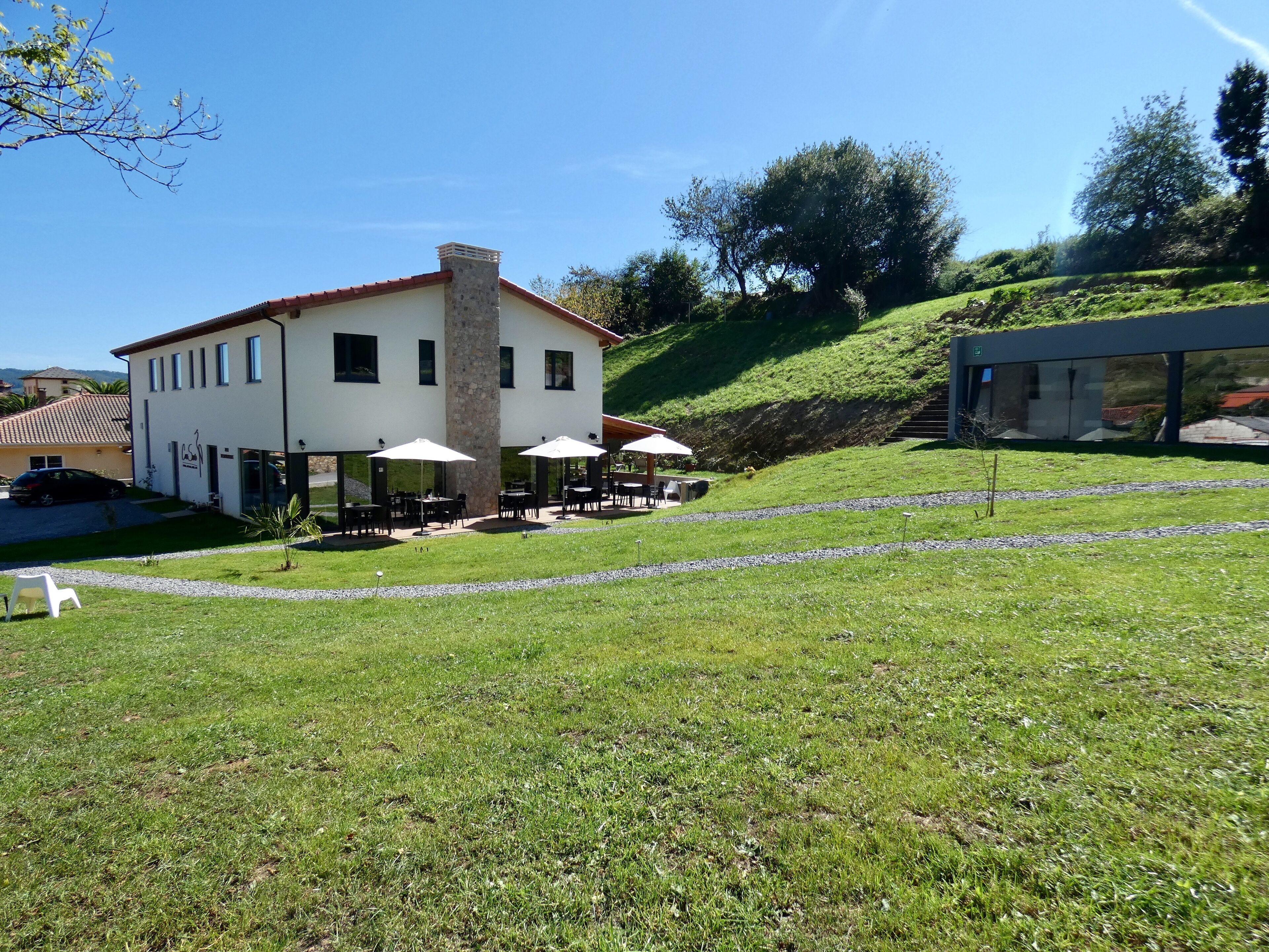 Un bâtiment à deux étages avec une cheminée en pierre, un espace extérieur aménagé avec des parasols et une colline verdoyante en arrière-plan.