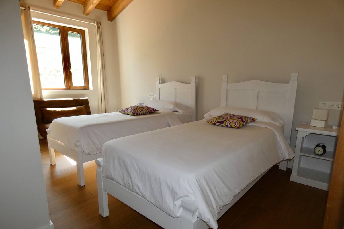 A bedroom with two single beds, a wooden floor, a nightstand with a clock, and a window with curtains.