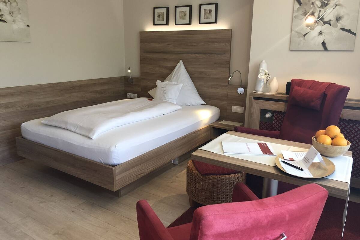 Ett hotellrum med en säng i trä, ett skrivbord med en stol, ett litet bord med en skål med frukt och en röd stol.