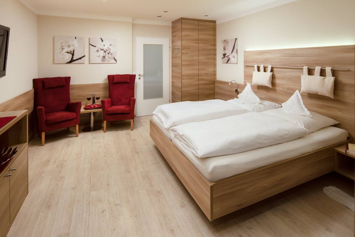 Ett modernt hotellrum med en säng i trä, två röda fåtöljer, ett litet bord med vinglas och konstverk på väggarna.