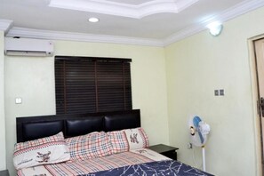 A bedroom with a bed, a window with blinds, an air conditioning unit, a bedside table, and a wall-mounted fan. - Unique Didis Royal Hotel & Suites (Lagos)