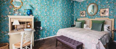 A bedroom with floral wallpaper, a wooden desk, a bed with green pillows, and a mirror above the bed.