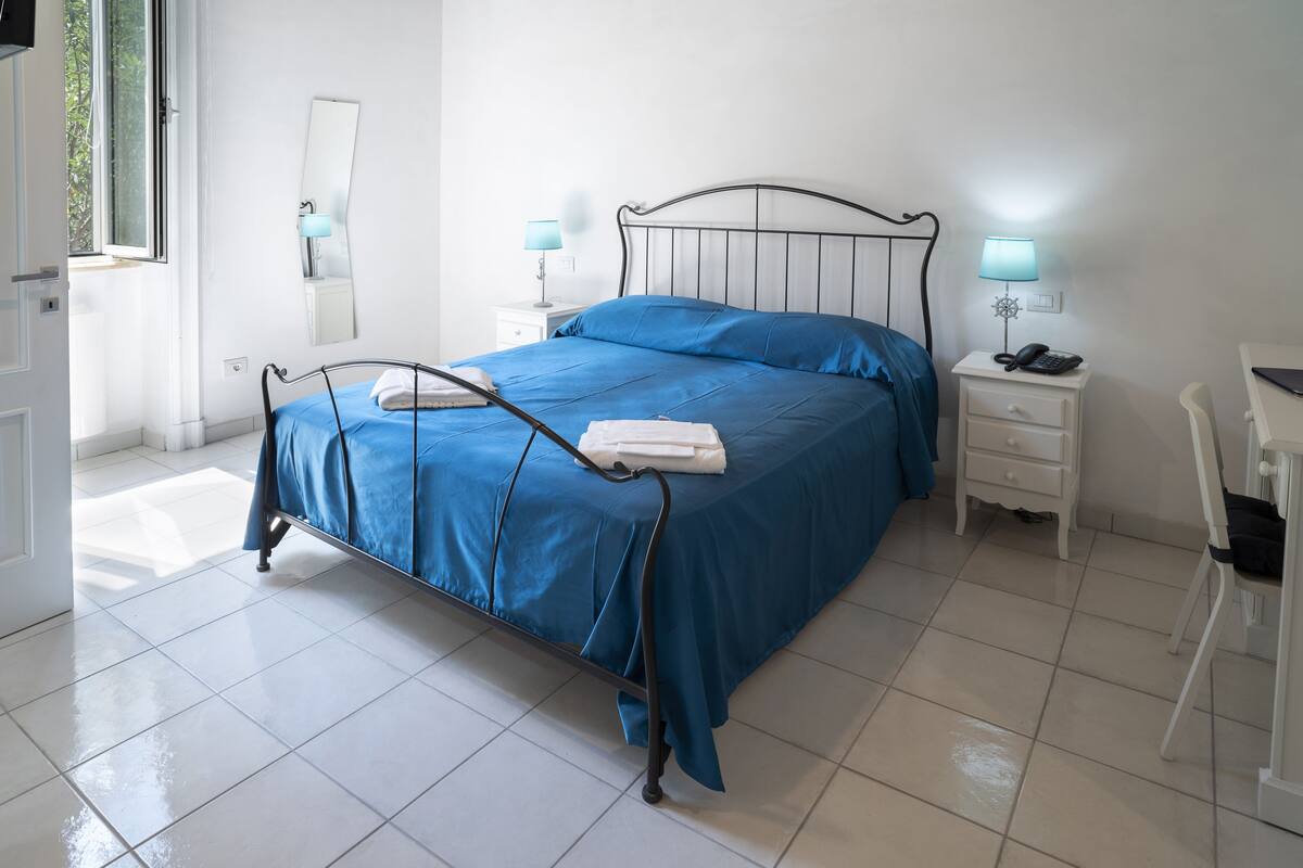 Una cama bien hecha con ropa de cama azul, una mesita de noche con lámpara y una silla.