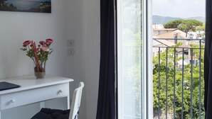 A room with a white desk, a vase with flowers, and a view of a balcony with greenery and buildings. - Residenza d'Epoca Olimpia (Castellabate)