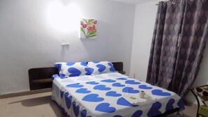 A single bed with a white and blue heart-patterned bedspread, a small painting on the wall, and a window with curtains. - Hotel Restaurant du 3ème Pont (Abidjan)