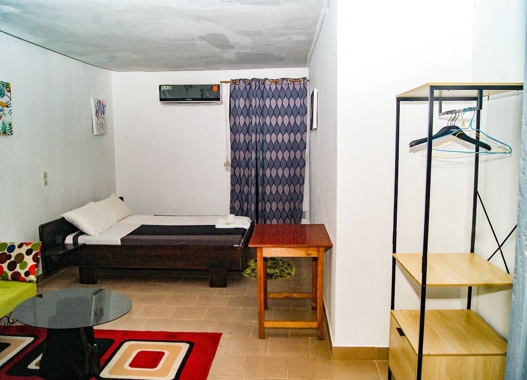 A small, compact living space with a bed, a small table, a hanging clothes rack, and a red rug.