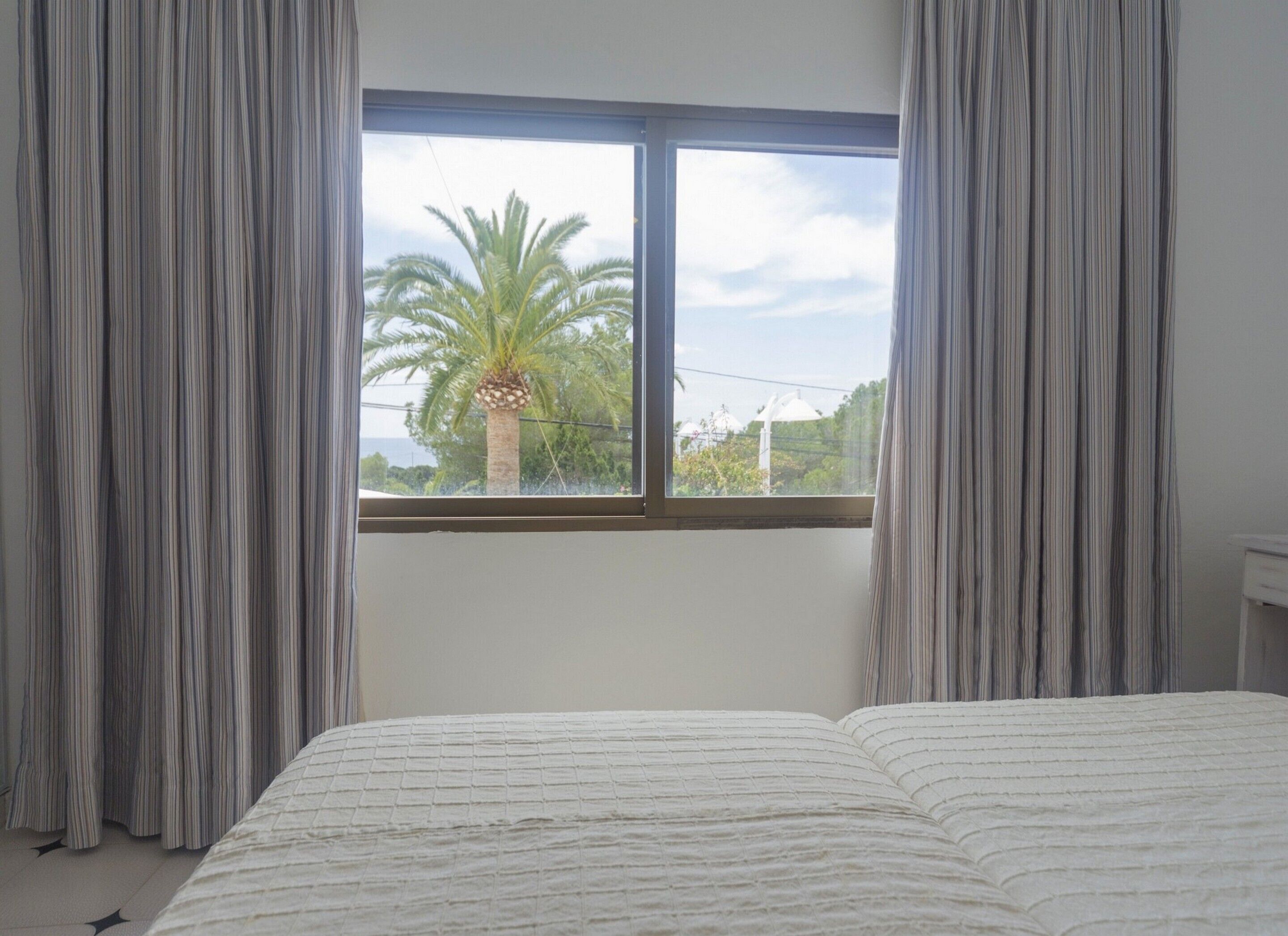 A bedroom with a large window, a bed with a white textured bedspread, and grey curtains.