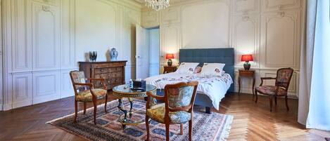 A bedroom with a large bed, two bedside tables with lamps, a chandelier, a wooden floor, and a patterned rug.