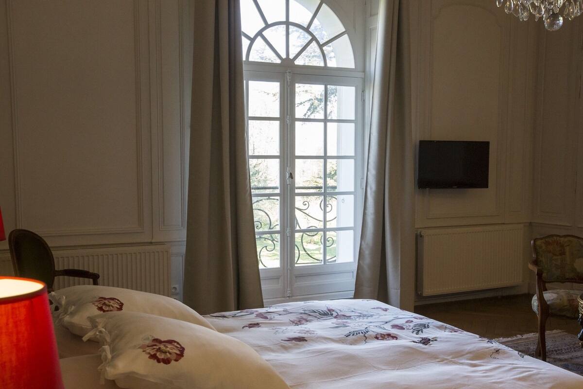 Ein Schlafzimmer mit großem Fenster, einem Bett mit floralem Bettbezug, einer roten Lampe und einem an der Wand befestigten Fernseher.