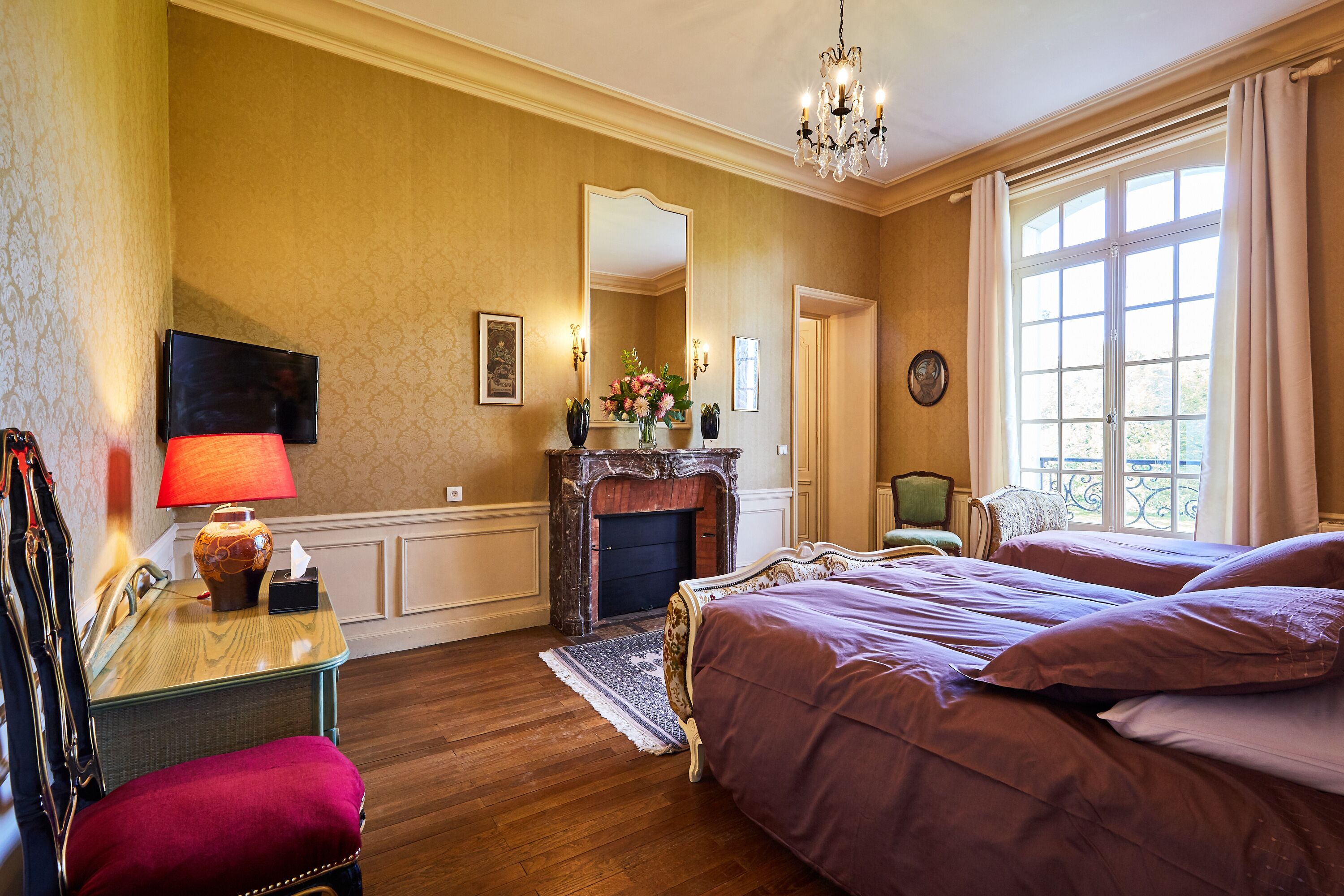 A hotel room with a wooden floor, a fireplace, a bed with purple bedding, a desk with a lamp, and a television on the wall.