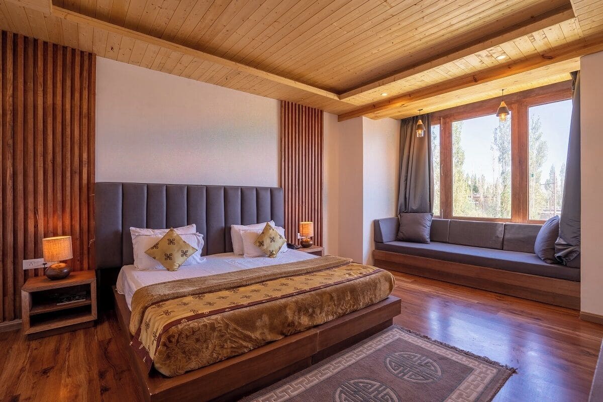 A bedroom with a large bed, a wooden headboard, a bench with cushions, and a window with curtains.