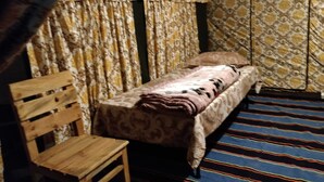 A small, single-bed room with a patterned bedspread, a wooden chair, and striped carpeting. - Camp Nirmit By Royal Collection (Dhari)