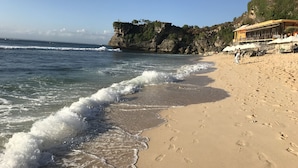 A beach with clear waters, a rocky cliff, and a building with outdoor seating. - Villa Viking (Jimbaran)