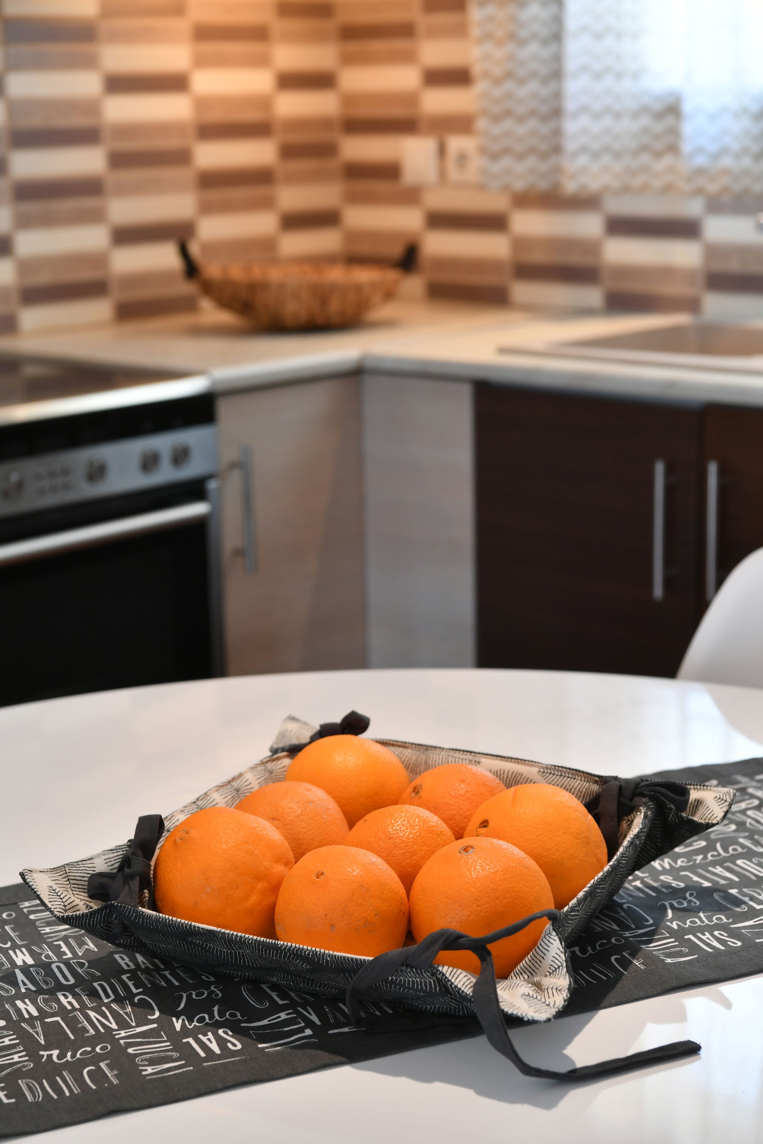A kitchen with a table holding a basket of oranges, a stove, and a tiled backsplash.