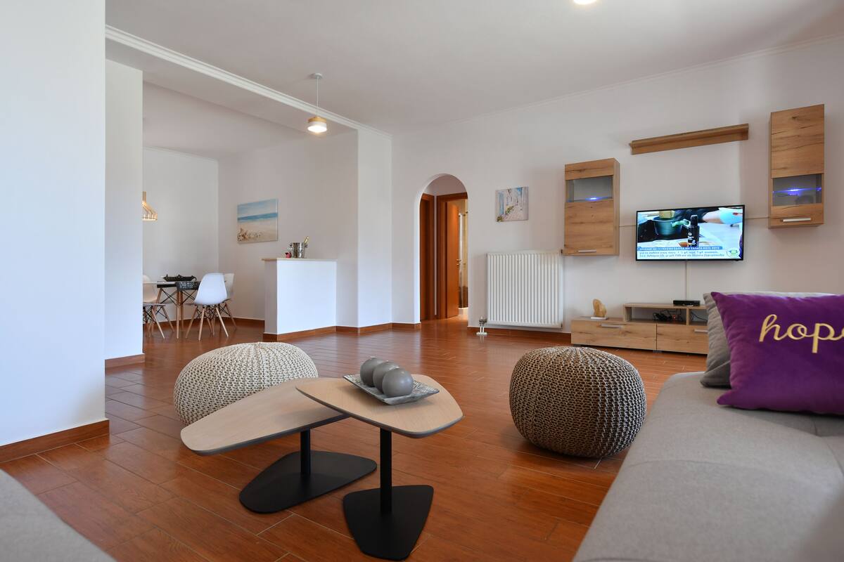 Una sala de estar moderna con un sofá, un reposapiés y una mesa con objetos decorativos. Hay un televisor montado en la pared y una zona de cocina al fondo.