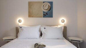 Un lit bien fait, avec une literie blanche, deux oreillers, un coussin décoratif orné du mot « HOME », un chapeau et des lunettes de soleil accrochés au mur, et deux tables de chevet avec des lampes murales.