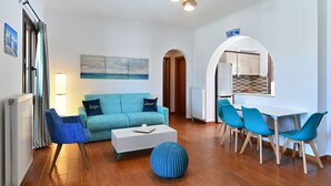 Un salon moderne avec un canapé, une table basse et des fauteuils, un coin repas avec une table et des chaises, et une cuisine visible à travers une arche.