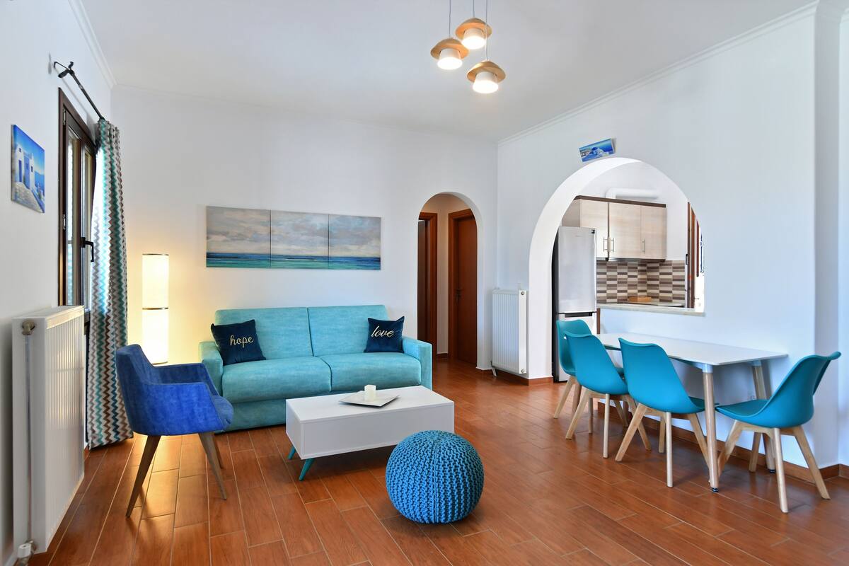 Una sala moderna con sofá, mesa de centro y sillones, un comedor con mesa y sillas, y una cocina visible a través de un arco.