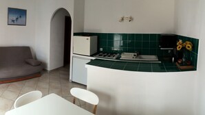 A compact kitchen with a gas stove, a small refrigerator, and a sink. There is a white table with two chairs, a grey sofa, and a painting on the wall. - Villa STELLA D ORO, F2 * (Porto-Vecchio)